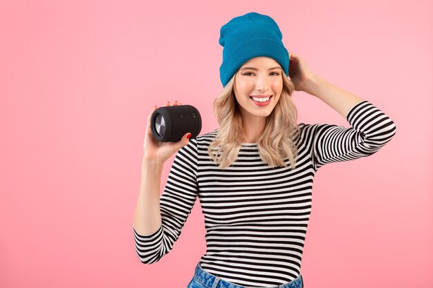 Mulher jovem e bonita segurando um alto-falante sem fio, ouvindo música, usando uma camisa listrada e chapéu azul, sorrindo, feliz humor positivo, posando na parede rosa