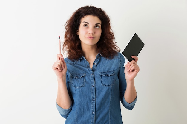 Mulher jovem e bonita segurando um caderno e um lápis, pensando, olhando para cima, tendo uma ideia, cabelo encaracolado, pensativo, isolado, camisa jeans azul, estilo hippie, aprendizagem do aluno, educação