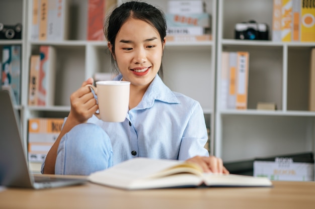 Mulher jovem e bonita sentada e usando o laptop e o livro didático para trabalhar ou aprender on-line, segurando a caneca de café na mão e sorrindo com felicidade