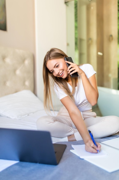 Mulher jovem e bonita sentada na cama com seu laptop e falando no telefone com um bloco de notas ao redor
