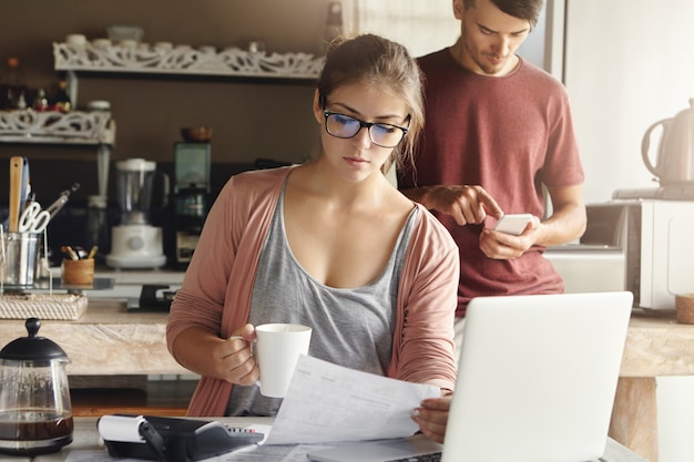 Mulher jovem e bonita séria em copos retangulares tomando café e estudando a conta na mão, sentado no interior da cozinha moderna na frente do laptop aberto