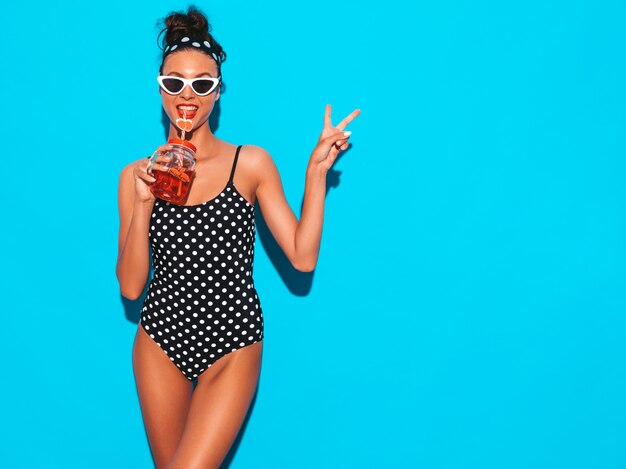Mulher jovem e bonita sexy hipster sorridente em óculos de sol. Garota de maiô de ervilhas de verão maiô. Posando perto da parede azul, bebendo bebida smoozy cocktail fresca. Mostra o sinal de paz