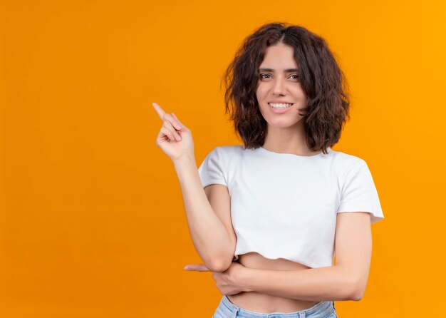 Mulher jovem e bonita sorridente apontando para o lado esquerdo em uma parede laranja isolada com espaço de cópia