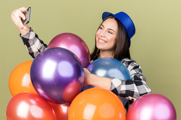 Mulher jovem e bonita sorridente com chapéu azul em pé atrás de balões tira uma selfie isolada na parede verde oliva