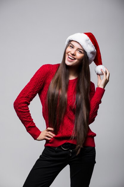 Mulher jovem e bonita sorridente com chapéu de Papai Noel em pé e olhando por cima da parede cinza