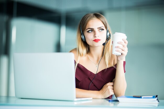 Mulher jovem e bonita sorridente com fones de ouvido e uma xícara de café olhando para o escritório