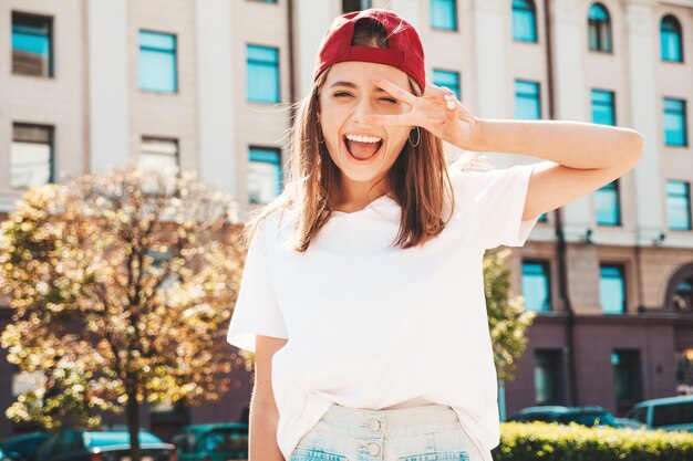 Mulher jovem e bonita sorridente hipster em camiseta branca de verão na moda Mulher despreocupada sexy posando no fundo da rua no boné ao pôr do sol Modelo positivo ao ar livre Alegre e feliz Mostra sinal de paz