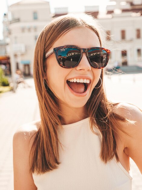 Mulher jovem e bonita sorridente hipster em roupas da moda de verão Mulher despreocupada sexy posando no fundo da rua ao pôr do sol Modelo positivo ao ar livre Alegre e feliz em óculos de sol