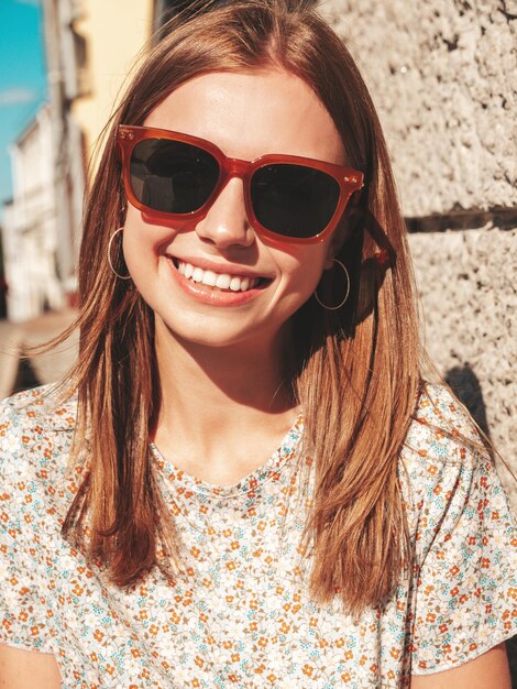 Mulher jovem e bonita sorridente hipster em roupas da moda de verão Mulher despreocupada sexy posando no fundo da rua ao pôr do sol Modelo positivo ao ar livre Alegre e feliz em óculos de sol