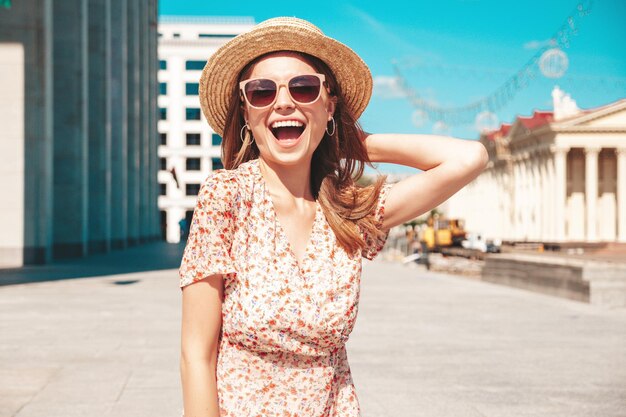 Mulher jovem e bonita sorridente hipster em roupas da moda de verão Mulher despreocupada sexy posando no fundo da rua ao pôr do sol Modelo positivo ao ar livre Alegre e feliz em óculos escuros e chapéu