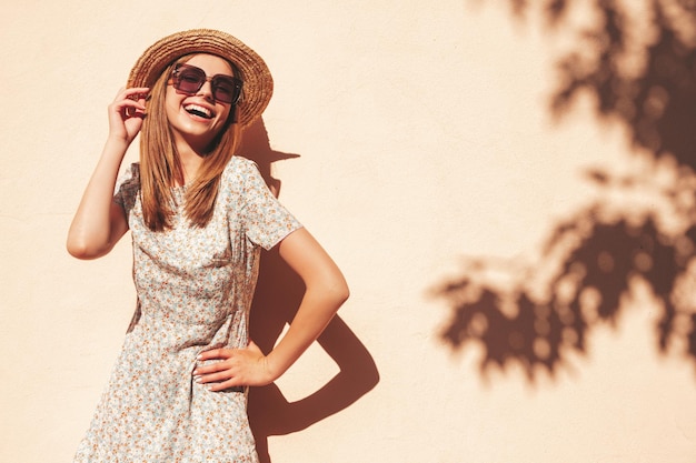 Mulher jovem e bonita sorridente hipster em vestido de verão na moda Mulher despreocupada sexy posando na rua perto da parede no chapéu ao pôr do sol Modelo positivo ao ar livre em óculos de sol Alegre e feliz