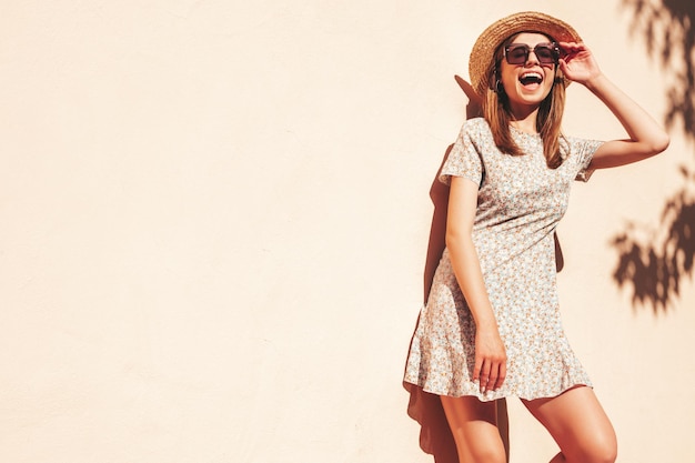 Foto grátis mulher jovem e bonita sorridente hipster em vestido de verão na moda mulher despreocupada sexy posando na rua perto da parede no chapéu ao pôr do sol modelo positivo ao ar livre em óculos de sol alegre e feliz