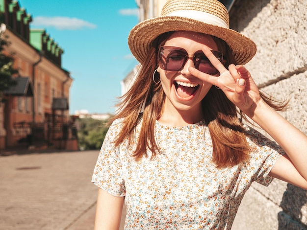 Foto grátis mulher jovem e bonita sorridente hipster em vestido de verão na moda mulher despreocupada sexy posando no fundo da rua no chapéu ao pôr do sol modelo positivo rindo ao ar livre alegre e feliz em óculos de sol