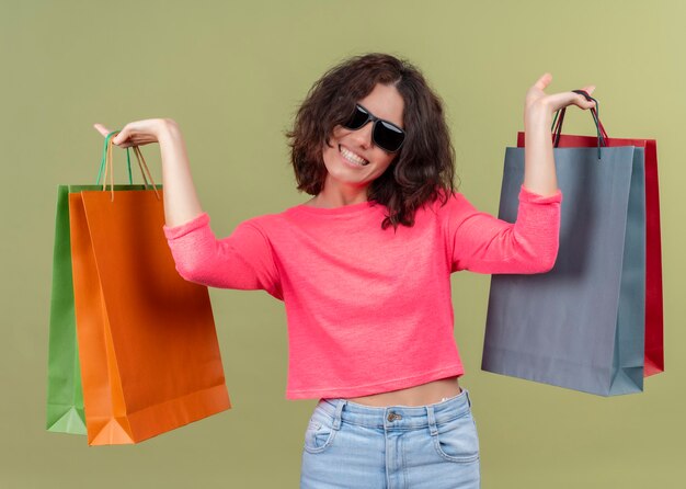 Mulher jovem e bonita sorridente levantando sacolas de papelão na parede verde isolada