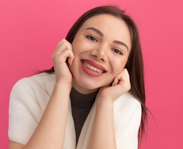 Mulher jovem e bonita sorridente mantendo as mãos nas bochechas isoladas na parede rosa