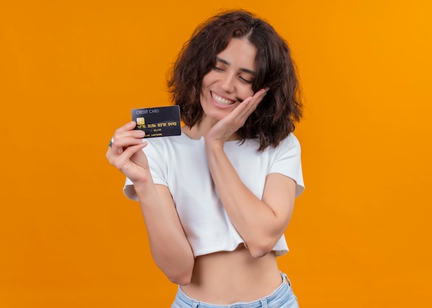 Mulher jovem e bonita sorridente segurando o cartão e colocando a mão no rosto em uma parede laranja isolada com espaço de cópia