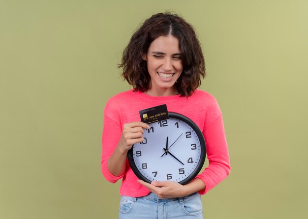 Mulher jovem e bonita sorridente segurando o cartão e o relógio na parede verde isolada com espaço de cópia