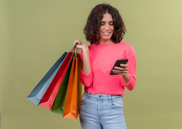 Foto grátis mulher jovem e bonita sorridente segurando sacolas de papelão e um telefone celular na parede verde isolada