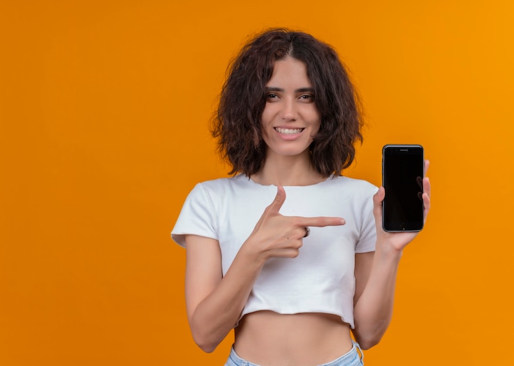 Foto grátis mulher jovem e bonita sorridente segurando um telefone celular e apontando com o dedo para ele na parede laranja isolada com espaço de cópia