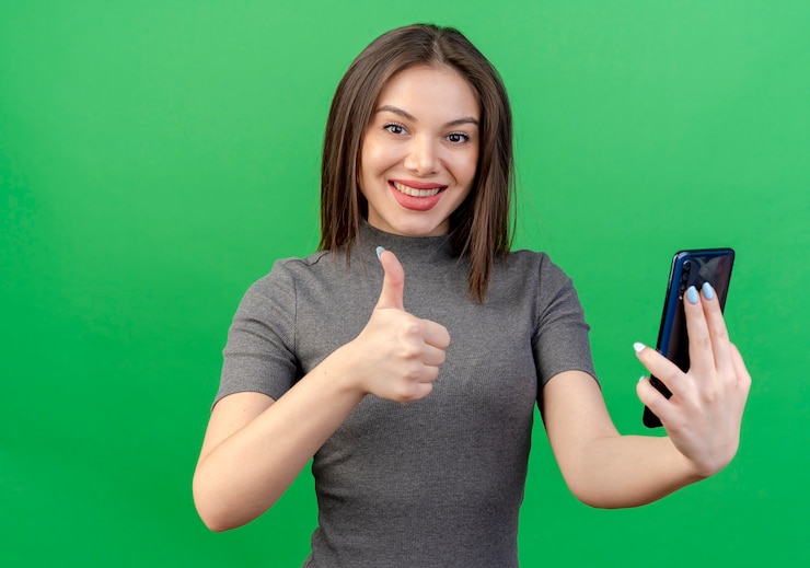 Foto grátis mulher jovem e bonita sorridente segurando um telefone celular e mostrando o polegar isolado no fundo verde