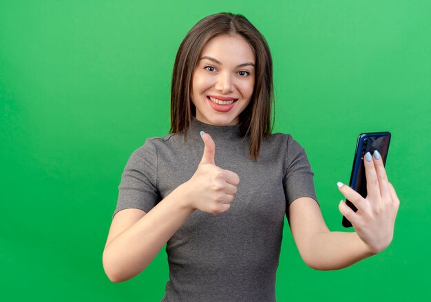 Mulher jovem e bonita sorridente segurando um telefone celular e mostrando o polegar isolado no fundo verde