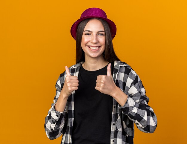 Mulher jovem e bonita sorridente usando chapéu de festa aparecendo os polegares isolados na parede laranja