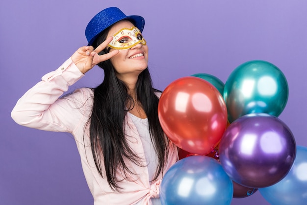 Mulher jovem e bonita sorridente usando chapéu de festa e máscara de baile de máscaras segurando balões, mostrando um gesto de paz isolado na parede azul