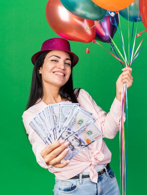 Mulher jovem e bonita sorridente usando chapéu de festa segurando balões e segurando dinheiro na frente, isolado na parede verde
