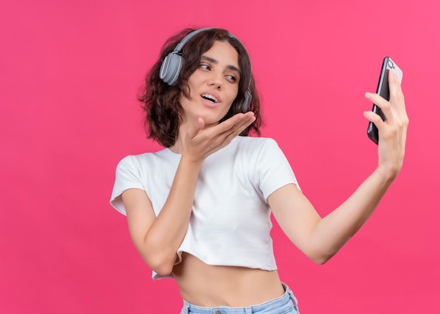 Mulher jovem e bonita sorridente, usando fones de ouvido e segurando o celular, mandando beijo na parede rosa com espaço de cópia