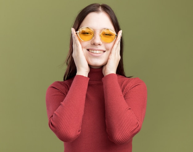 Mulher jovem e bonita sorridente usando óculos escuros, com as mãos no rosto, olhando para a frente, isolada na parede verde oliva