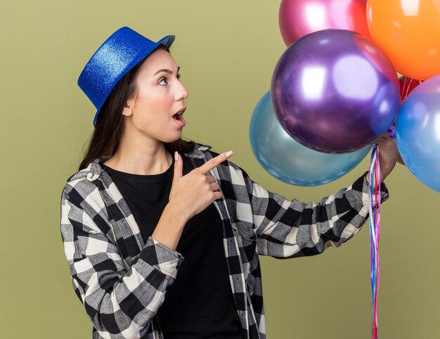 Mulher jovem e bonita surpresa com chapéu azul segurando e apontando para balões isolados na parede verde oliva