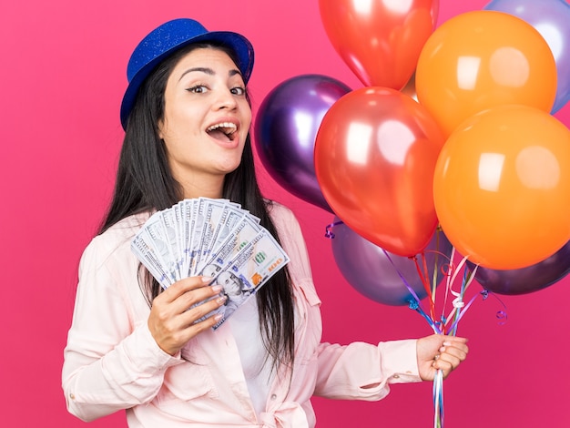 Mulher jovem e bonita surpresa com chapéu de festa segurando balões com dinheiro isolado na parede rosa