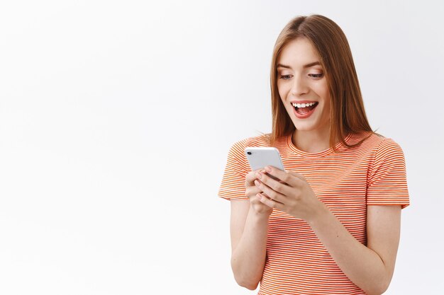 Mulher jovem e bonita surpresa e entusiasmada em uma camiseta listrada, segurando o smartphone lendo notícias incríveis, boca aberta fascinada, sorrindo reagindo aos preços maravilhosos no site de compras de aplicativos para dispositivos móveis