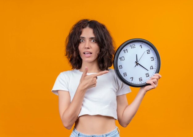 Foto grátis mulher jovem e bonita surpresa segurando o relógio e apontando com o dedo para ele na parede laranja isolada com espaço de cópia