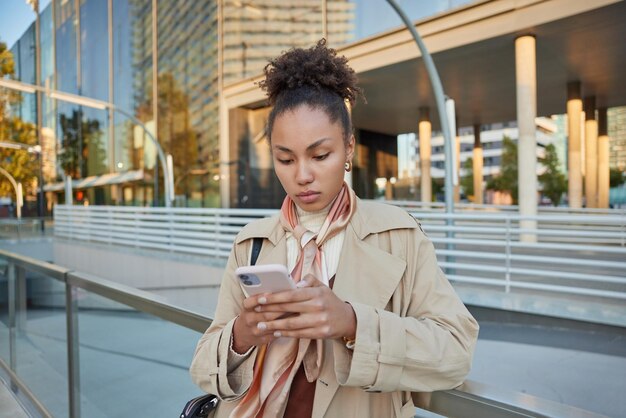 Mulher jovem e bonita tem expressão séria usa celular verifica bate-papos de notificação on-line via aplicativo satisfeito com boa conexão de internet assiste poses de vídeo contra ambiente urbano