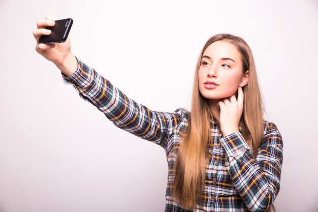 Mulher jovem e bonita tirando foto de selfie com smartphone isolado na parede branca