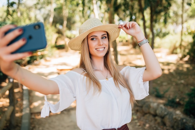 Mulher jovem e bonita tomando uma selfie com um chapéu elegante de verão em um parque.