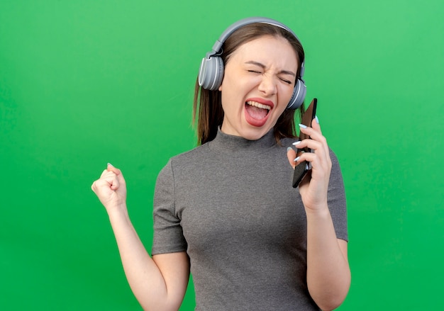 Mulher jovem e bonita usando fones de ouvido cantando com os olhos fechados e os punhos cerrados e usando o celular como microfone isolado sobre fundo verde