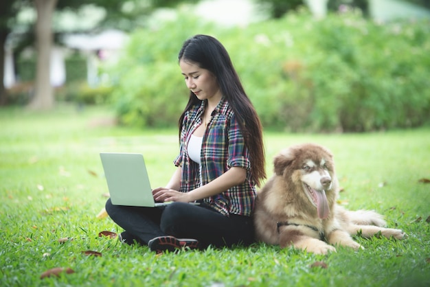 Mulher jovem e bonita usando laptop com seu cachorro pequeno em um parque ao ar livre. Retrato do estilo de vida.