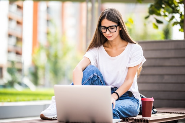 Mulher jovem e bonita usando laptop enquanto está sentado em um banco, bebendo a xícara de café para viagem