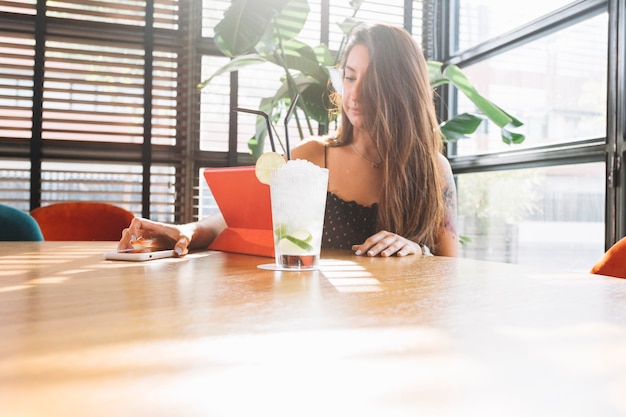 Foto grátis mulher jovem e bonita usando telefone celular com copo de coquetel na mesa de madeira