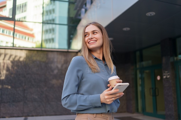 Mulher jovem e bonita usando um aplicativo em seu smartphone para enviar uma mensagem de texto perto de edifícios comerciais