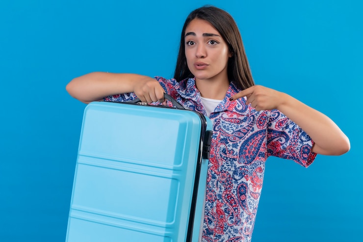 Foto grátis mulher jovem e bonita viajante segurando uma mala azul apontando com o dedo para ela, parecendo confusa e surpresa de pé sobre um fundo azul