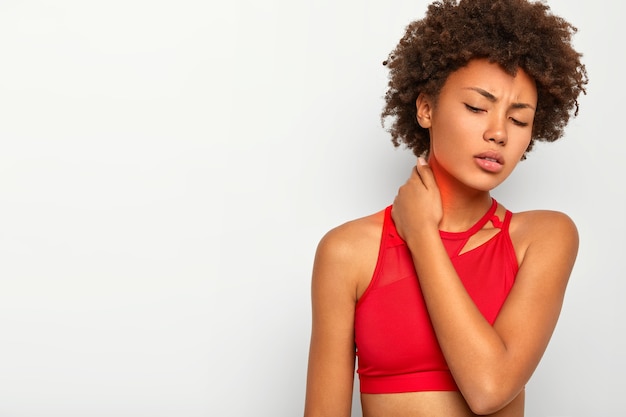 Foto grátis mulher jovem e cacheada cansada toca o pescoço, mantém os olhos fechados, usa uma blusa vermelha, inclina a cabeça, estando exausta, posa contra um fundo branco para seu conteúdo promocional