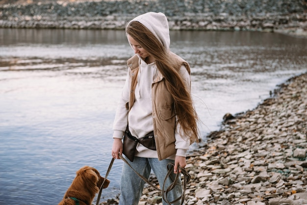 Mulher jovem e cão retriever caminham na margem do rio na temporada de outono