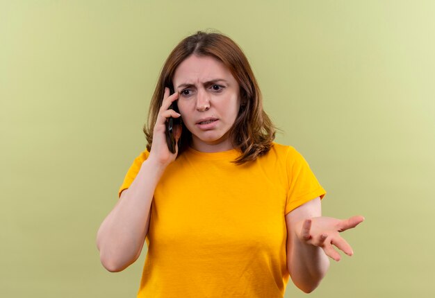 Mulher jovem e casual descontente falando no telefone e mostrando a mão vazia na parede verde isolada