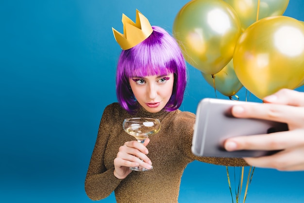 Mulher jovem e charmosa com corte de cabelo roxo, coroa na cabeça fazendo retrato de selfie. balões dourados, champanhe, festa de ano novo, vestido de luxo, maquiagem de ouropel.