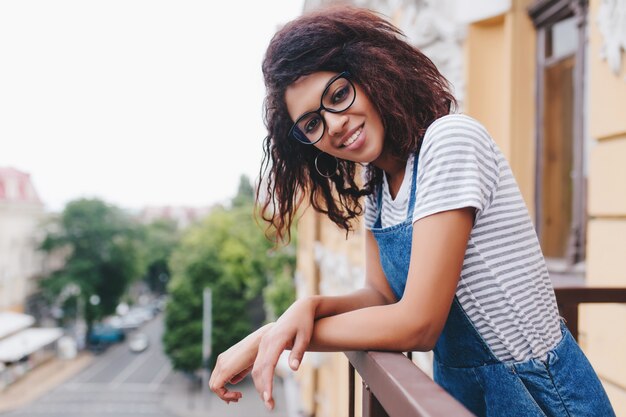 Mulher jovem e charmosa de óculos posando em pé no terraço e apreciando a vista da cidade