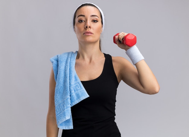 Mulher jovem e confiante, bonita e esportiva, usando bandana e pulseiras segurando halteres com uma toalha no ombro, olhando para a frente, isolada na parede branca com espaço de cópia