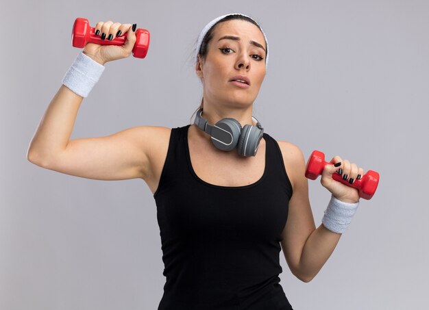 Mulher jovem e confiante, bonita e esportiva, usando fita para a cabeça e pulseiras levantando halteres com fones de ouvido ao redor do pescoço, olhando para a frente, isolado na parede branca